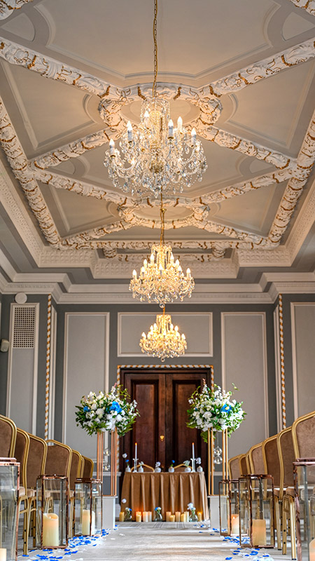 Floor-level view of central aisle with floral domes on copper stands, copper lanterns with warm LED candlelight, and grand chandeliers in the museum ceremony room at Manchester Hall