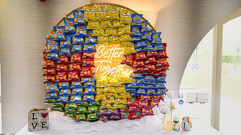 Mesh wall display of assorted crisps surrounding a neon ‘Better Together’ sign at a wedding reception in Merrydale Manor