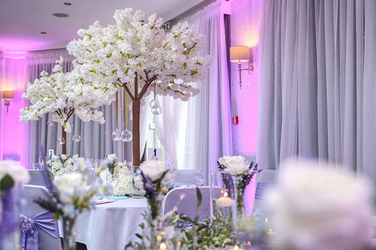 Tall white blossom tree table centrepieces in a wedding venue with elegant draping and candlelit décor.